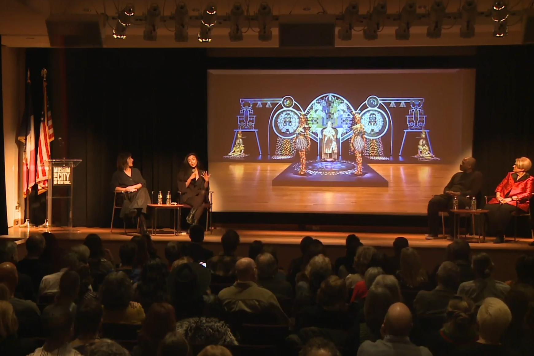 "Panel discussion at the Museum of the City of New York, featuring four speakers on stage with a projected artwork behind them, and an audience in attendance."